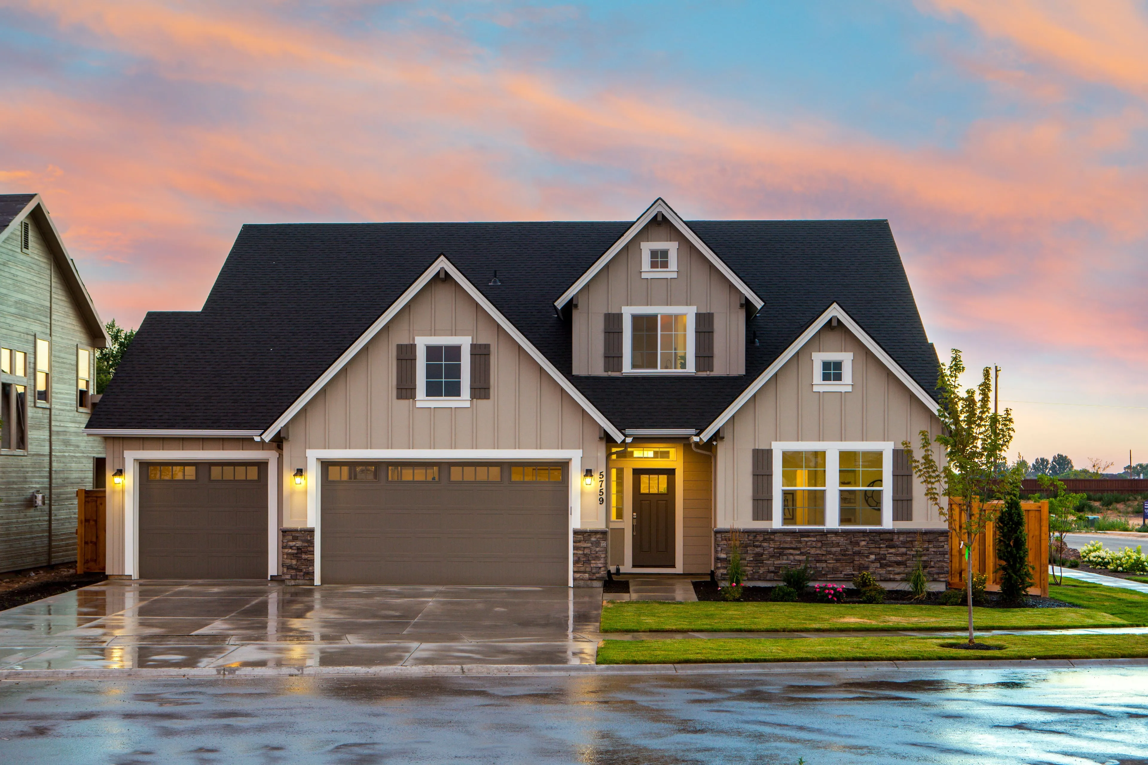 Two-story craftsman home at twilight, porch and garage lights on, wet driveway reflecting the sky.